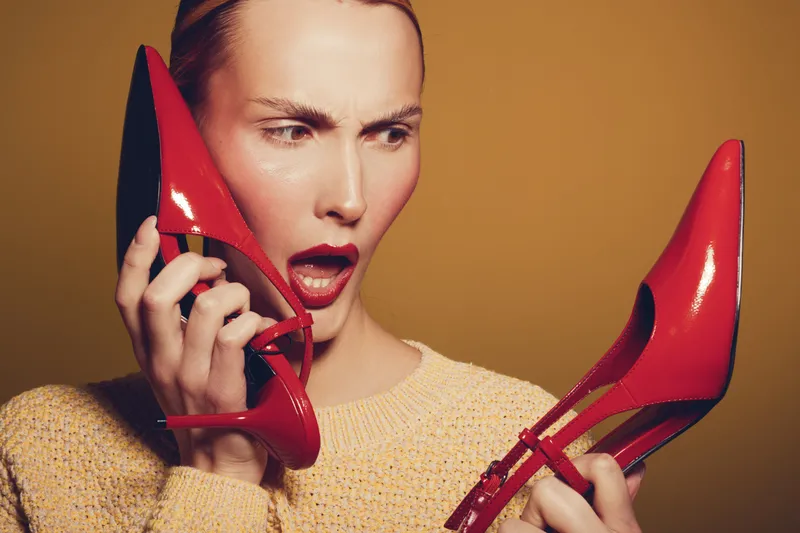 Surprised woman in yellow sweater holds red high heel shoes near face, expressing shock and curiosity. Studio portrait with warm brown background, concept of fashion and emotions.