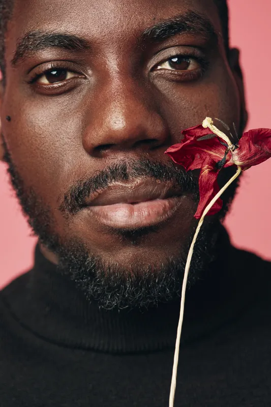 African man with dark skin and beard holding dried red flower close to face in creative conceptual visual style on pink background. Portrait with artistic expression and natural texture.