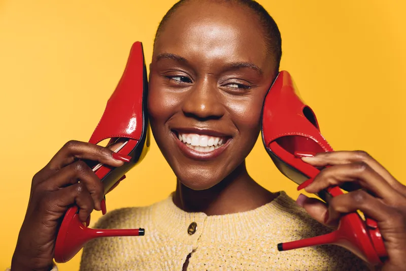 Smiling dark skinned African woman holds red high heels near her face against yellow background. Concept of fashion, beauty, style, and joyful emotions in studio lighting.