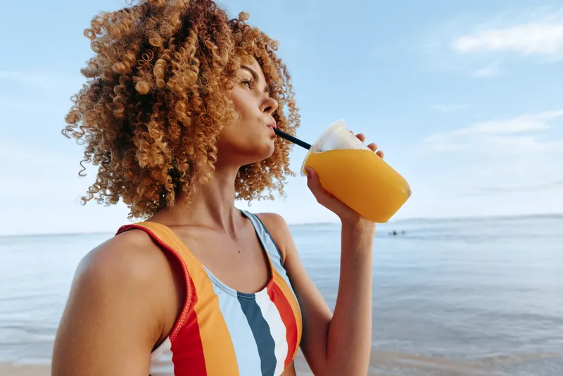 Woman with curly hair on a sunny beach drinks orange juice from a bottle, wearing a colorful swimsuit, enjoying a refreshing moment by the sea and bright sky