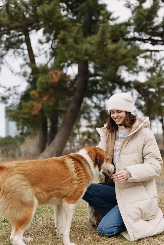 Woman kneels outdoors in a warm coat, giving gentle affection to a large dog as they share a calm moment in a park with pine trees, soft light, and a cozy winter touch.