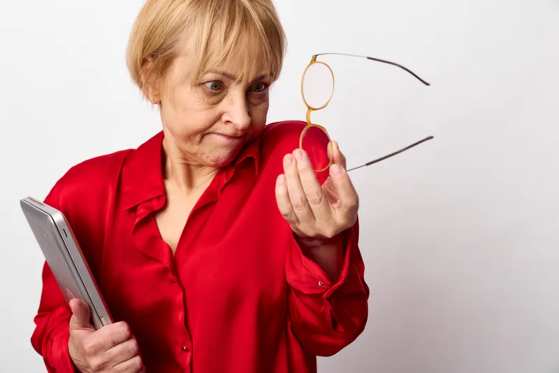 Elderly woman red shirt holding glasses and smartphone, vision concern in an indoor portrait against white background, attentive hands gesture, soft studio light