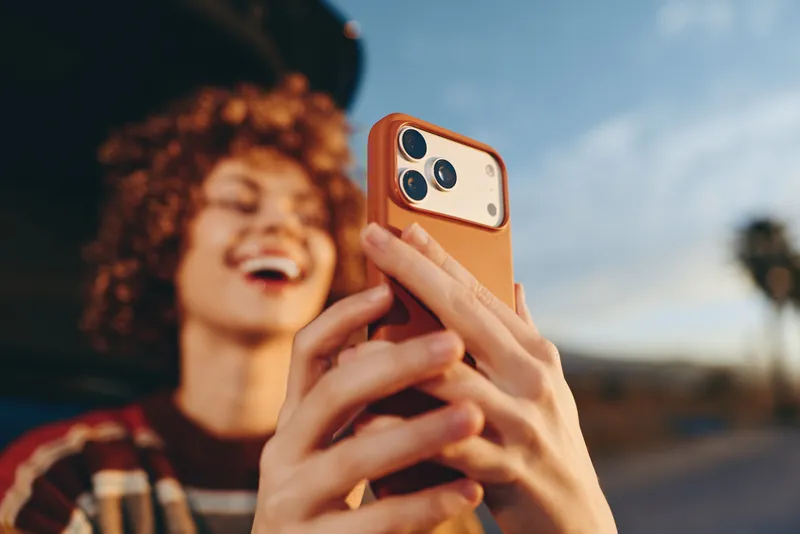 Smiling woman holding smartphone for a selfie during outdoors lifestyle moment, curly hair visible, orange phone case, blue sky and landscape softly blurred, candid portrait, relaxed