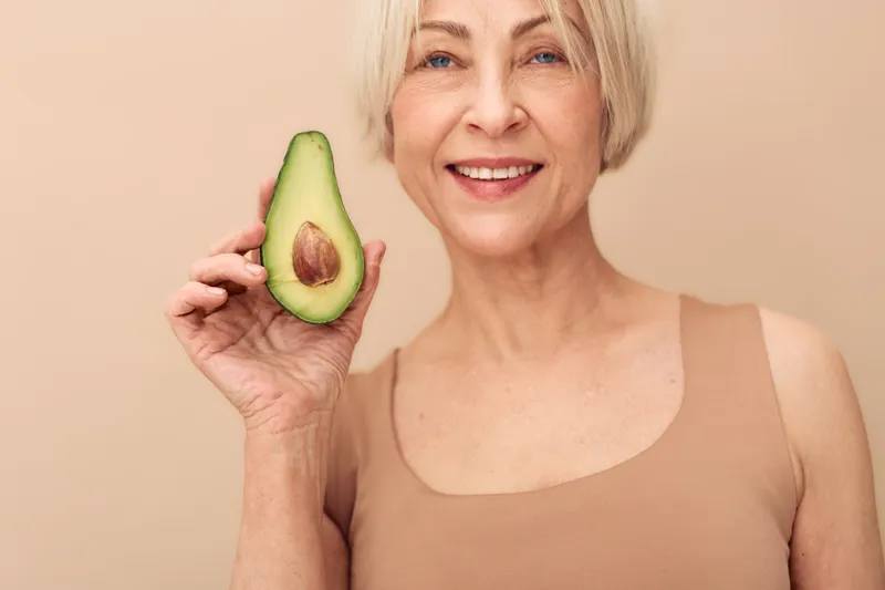 Smiling mature woman with short blonde hair holds half an avocado close to her face. Beige background and clothing create a natural, healthy lifestyle concept with fresh fruit and wellness.