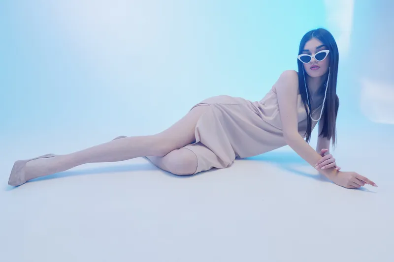 Young woman in a beige dress and white sunglasses lies on a light blue floor. Her long hair is straight, styled for a fashion shoot. Concept of beauty, fashion, and elegance.