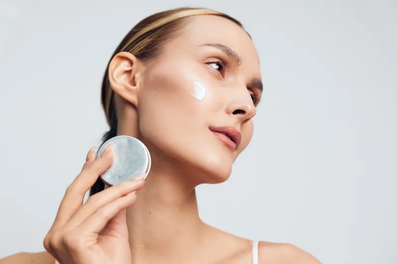 Young woman with clear skin applies moisturizer cream on her face using a cotton pad in a bright studio with a neutral background.