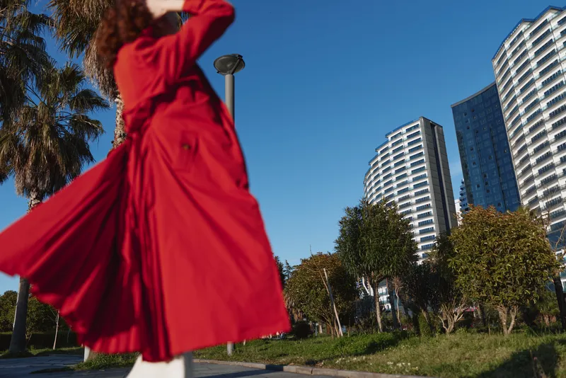 A woman in a flowing red coat walks outdoors near modern high-rise buildings under a clear blue sky. Green trees and palm trees are visible in the urban park setting.