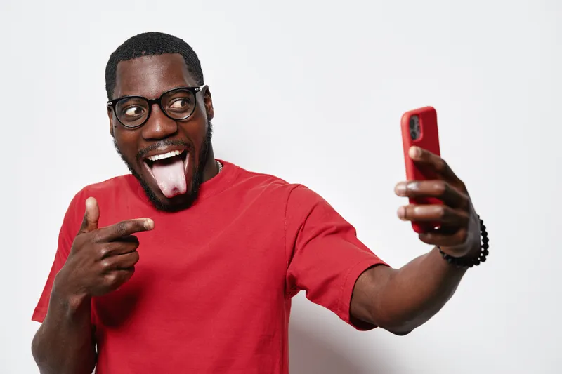 African American man with dark skin wearing glasses and red shirt taking selfie with smartphone, smiling and making playful face on white background in studio.