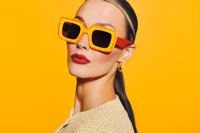 Stylish woman with slicked hair wears oversized orange sunglasses and red lipstick against bright yellow background. Concept of fashion, summer style, and bold accessories.