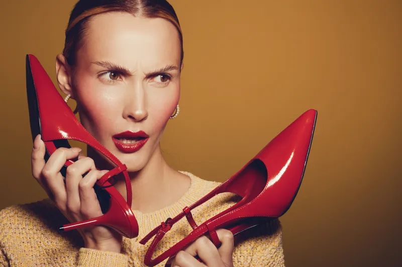 Confused woman with red lipstick holds two shiny red high heel shoes near her face on yellow background. Concept of fashion choice, style dilemma, and footwear comparison.
