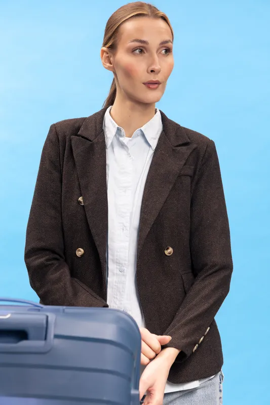 Young woman in brown blazer and white shirt stands with blue suitcase on blue background. Concept for travel, business trip, and professional journey with confident expression.