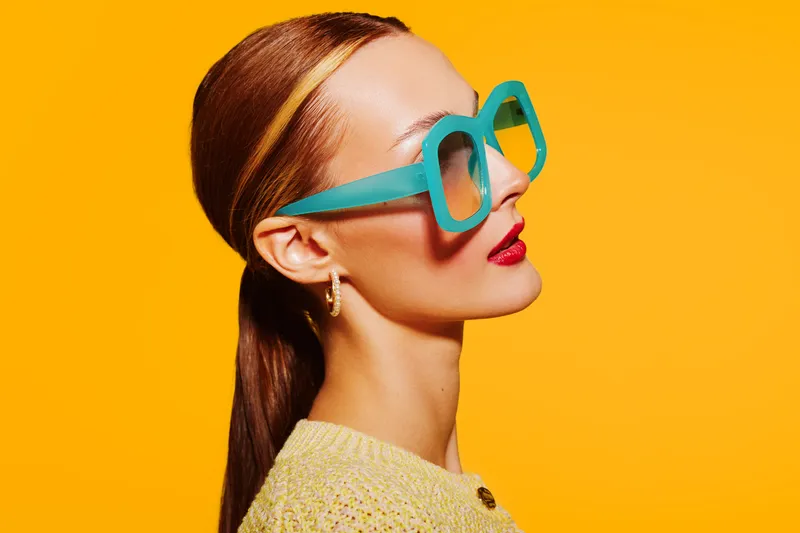 Profile of a stylish woman with slicked hair wearing large turquoise sunglasses and red lipstick against a bright yellow background. Concept of fashion, beauty, and summer style.