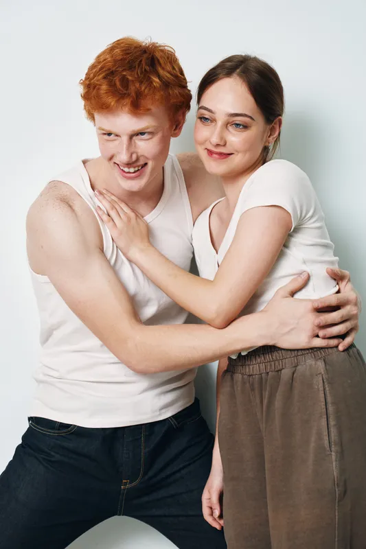 Couple smiling and hugging in casual clothes against white background. Young man and woman with happy facial expressions showing affection and closeness indoors.