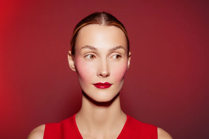 Young woman with red lipstick and flushed cheeks looks sideways against red background. Portrait of female model with smooth skin and sleek hair in red outfit. Beauty and fashion concept.