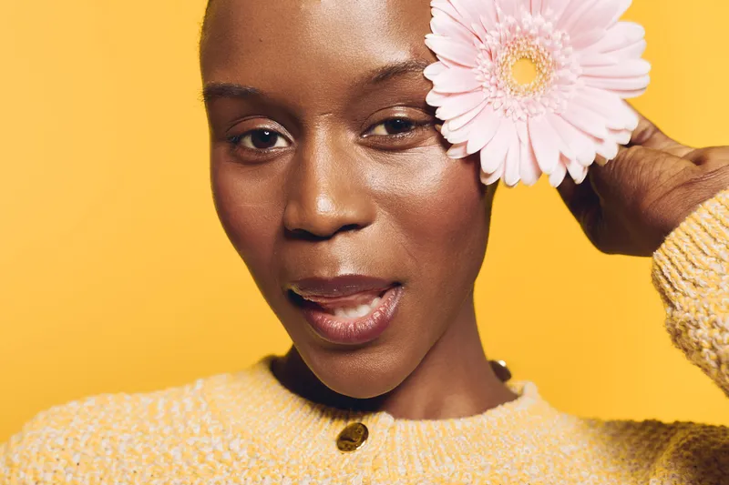 Close-up of a smiling black woman with dark skin holding a pink flower near her face. She wears a yellow sweater and poses against a bright yellow background. Concept of beauty and nature.