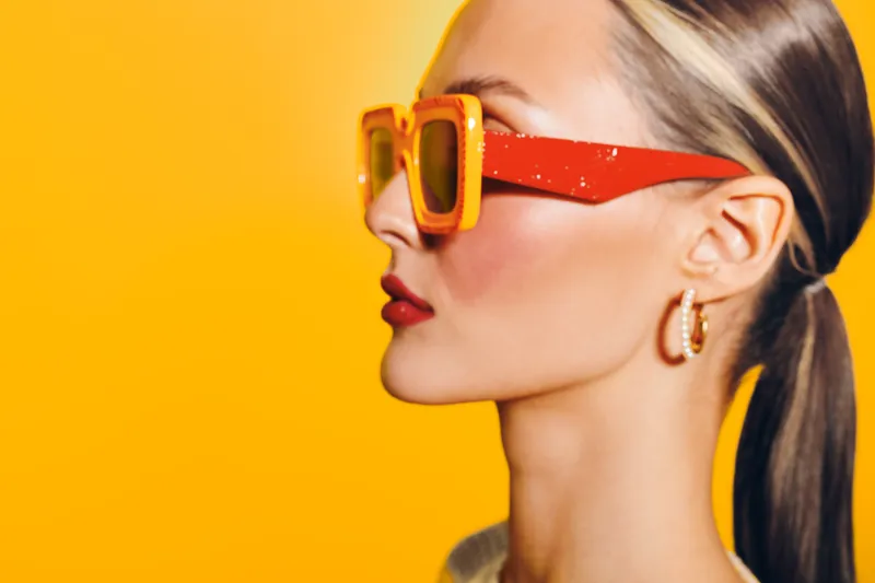 Blonde woman with ponytail wearing orange sunglasses and hoop earrings poses against bright yellow background. Fashion portrait with vibrant colors and stylish accessories concept.