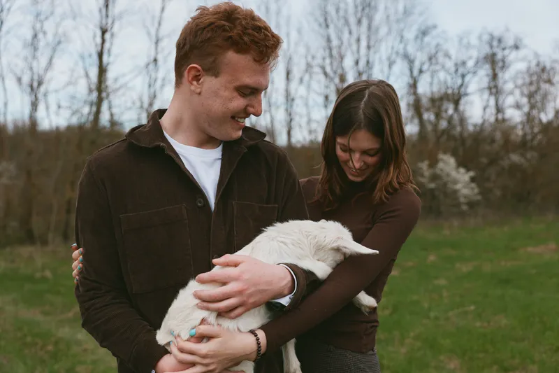 A young couple embraces outdoors while holding a white baby goat in their arms during a casual lifestyle moment in early spring.