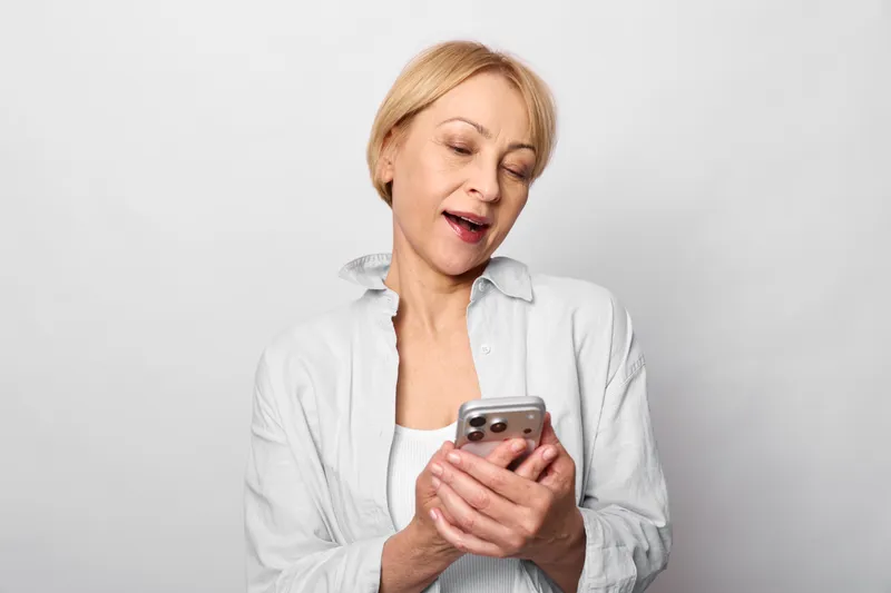 Woman holding smartphone looks upset, touching neck, studio portrait, light gray background, concerned about health symptoms, soft lighting shows stress and communication