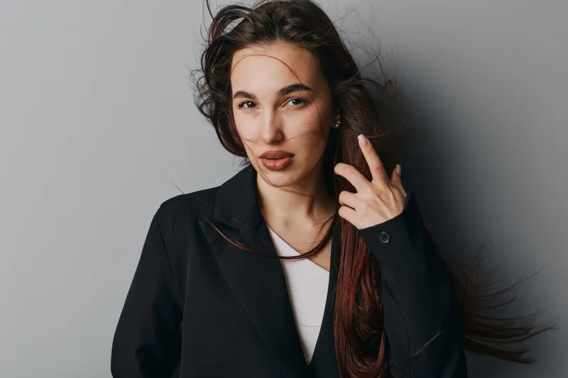 Young woman with long hair wearing black jacket posing confidently against gray background. Female model with natural makeup and serious expression in studio portrait photography.