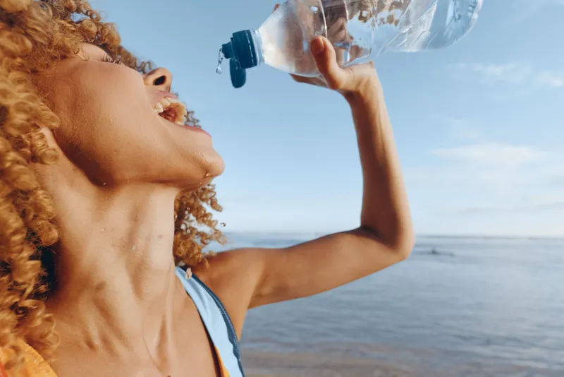Young woman with curly hair drinks water from a bottle at the beach, enjoying outdoor hydration under a sunny sky, sea in the background, capturing an active summer moment