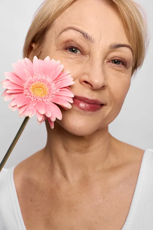 Mature woman portrait, closeup on smiling face, pink flower by cheek, soft studio light, fresh beauty mood, smooth skin, white background, skincare care