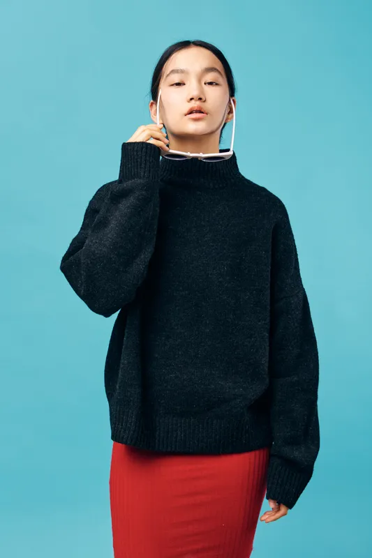 Young woman wearing black sweater and red skirt posing with white sunglasses on blue background. Fashion model looking confident with casual modern style and minimal makeup for portrait.