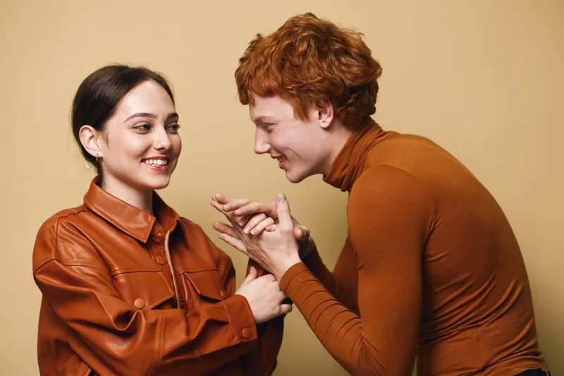 Young man and woman in casual clothing smiling and interacting together in studio. Friendly gesture and happy expression with warm brown tones on beige background.