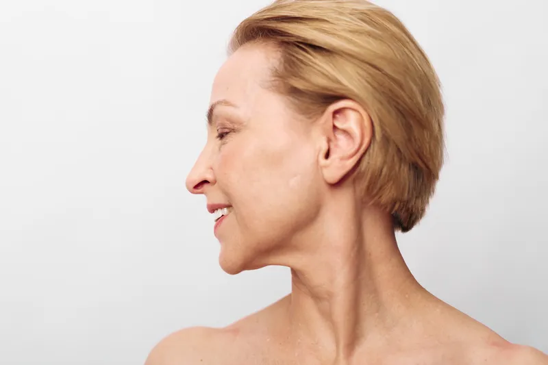 Profile of smiling mature woman with short blonde hair and clear skin on white background. Concept of natural beauty, aging gracefully, skincare and confident middle age.