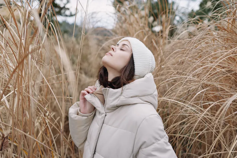 Woman in a plush winter coat and knit hat stands among tall grasses, gazing upward in an outdoor portrait, highlighting serene textures and natural calm.