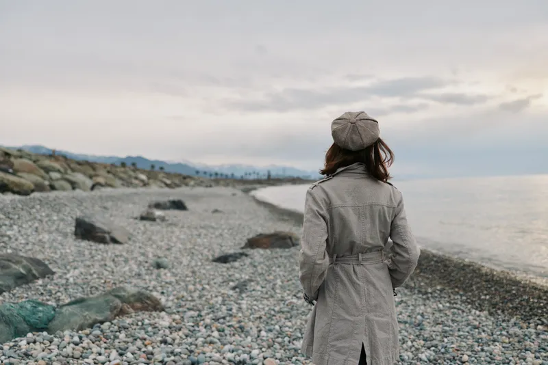 Woman in grey coat and hat stands on rocky beach looking at calm sea under cloudy sky. Concept for solitude, nature, autumn, and peaceful outdoor moment.
