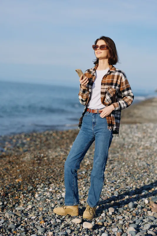 Beachside stroll by the sea with a casual dressed woman wearing a plaid shirt, jeans and sunglasses, enjoying a sunny day on a pebbled shoreline with relaxed confidence.
