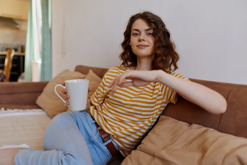 Young woman with curly brown hair sits on a cozy sofa at home, holding a mug and wearing a striped top, creating a relaxed mood of casual self care.