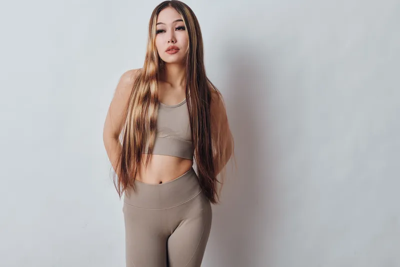 Young woman with long hair posing in beige sportswear against plain background, fitness and fashion concept. Confident female model with natural makeup and casual activewear outfit.