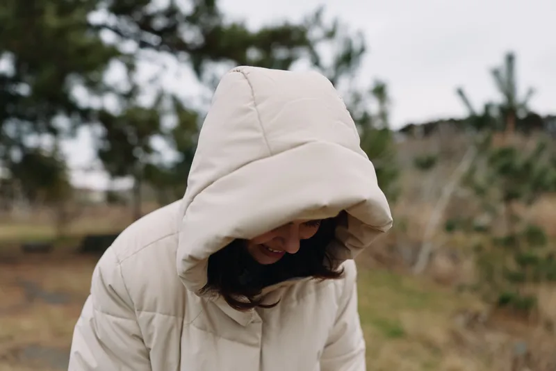 Hooded coat worn by a person in an outdoor setting, overcast day, embracing chilly weather with a tucked head and relaxed posture amid a natural landscape.