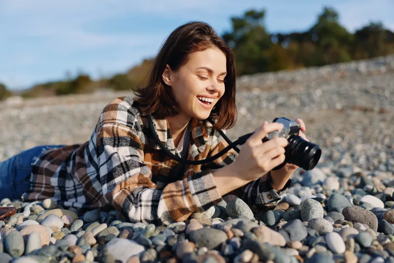 Woman photographer lying on a pebble beach, smiling as she holds a camera. Casual plaid shirt, sunny day, creative moment in nature, outdoor hobby photography and relaxed mood.