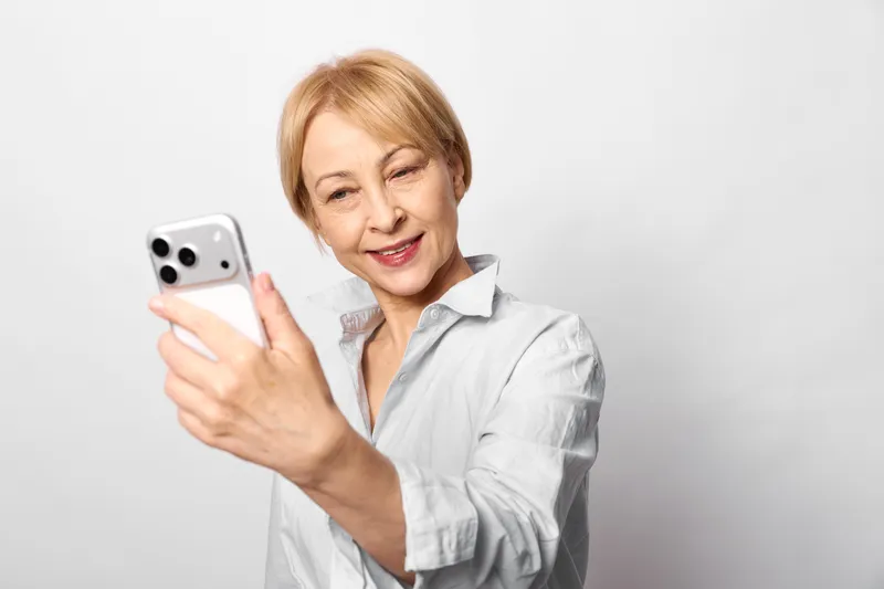 Elderly woman using smartphone for selfie, smiling in studio portrait with clean white background, showing digital communication and technology, calm mood and modern lifestyle today