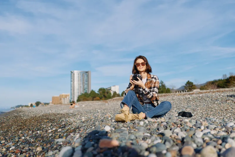 Woman on a pebble beach with a camera, casual attire and sunglasses, seated near the shore with a relaxed pose, urban buildings in the distance and a bright blue sky overhead.