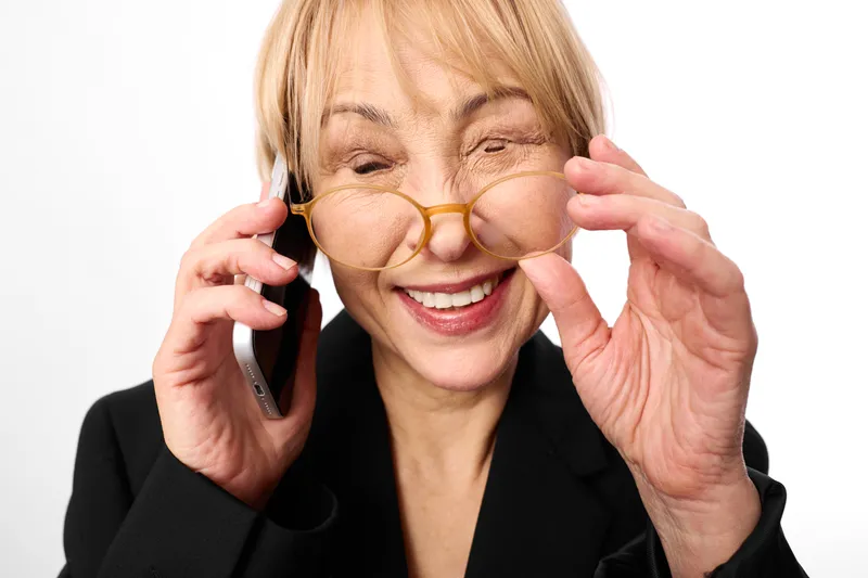 Senior woman with glasses holding smartphone and phone near ear, communicates with smile in business suit, indoor portrait on white background tech call today