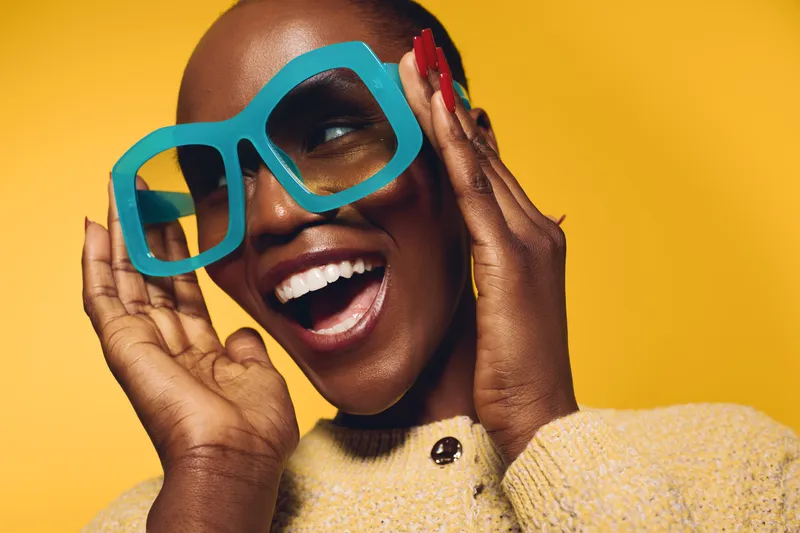 Happy dark skinned African American woman with short hair wearing large blue glasses and yellow sweater smiles joyfully on bright yellow background. Concept of fashion, style, and positivity.