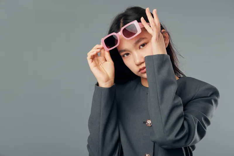Young woman with long dark hair wearing oversized gray blazer holding pink sunglasses near face with serious expression on simple gray studio background in fashion portrait.