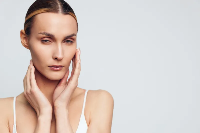 Calm closeup of a woman touching her face, clear skin and minimal makeup, soft studio lighting, white background, introspective mood, natural tones and clean aesthetic
