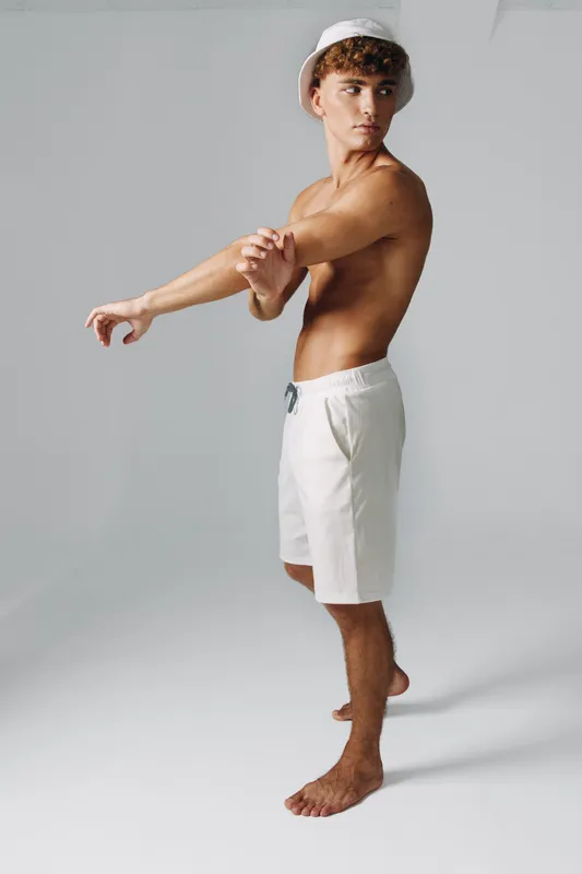 Young man in white shorts and bucket hat stretching his arm, standing barefoot on gray background, fitness and casual summer style with focused expression and toned body.