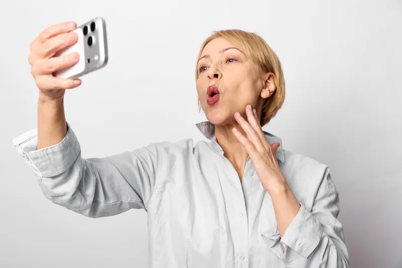 Woman selfie with smartphone, surprised portrait indoor, white closeup background, hand gesture with open mouth and raised fingers, soft light