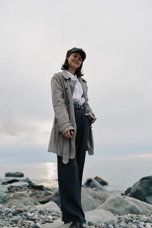 Young woman wearing a hat and long coat stands on rocky beach near ocean under cloudy sky. Casual fashion, outdoor lifestyle and autumn mood concept at coastline scene.