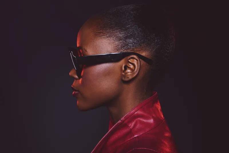 Profile of a confident black woman with dark skin wearing stylish glasses and a red leather jacket. Studio portrait with dramatic lighting on black background. Concept of fashion and empowerment.