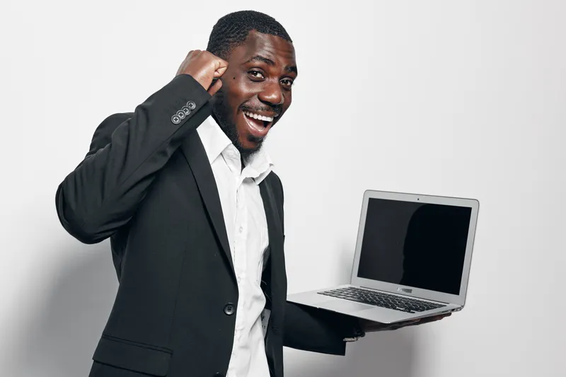 African American man with dark skin in black suit holding laptop and celebrating success with fist pump and smile on white background. Business, technology and achievement concept.