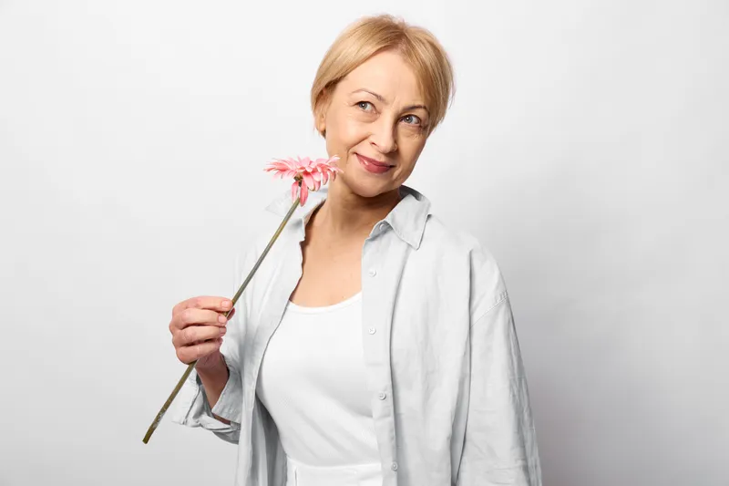 Woman senior smiling flower pink studio portrait holding blonde hair, gentle cheerful mood, mature casual shirt, holding pink blossom, soft studio light, white background