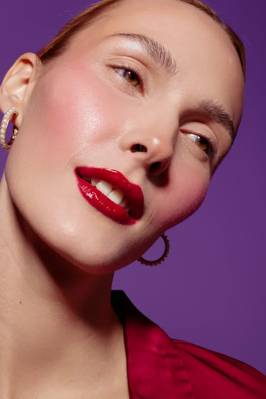 Close-up of a woman with glossy red lips and natural makeup wearing hoop earrings and a red blouse. Studio portrait on purple background with soft lighting and smooth skin texture.