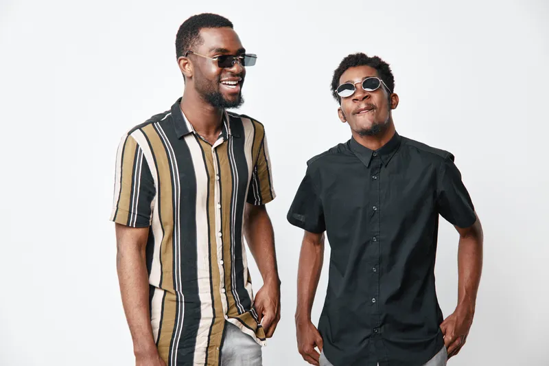Two young black men with dark skin wearing sunglasses and casual shirts standing and smiling on white background. African ethnicity, friendship and style with confident expressions.