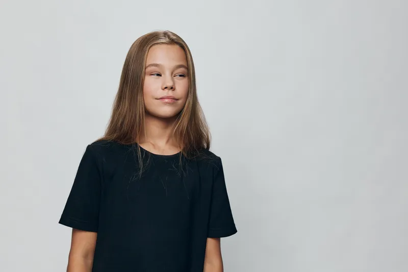 Girl with long hair wearing black shirt looks sideways with calm expression on light gray background. Studio portrait of thoughtful child with loose hair and casual outfit.